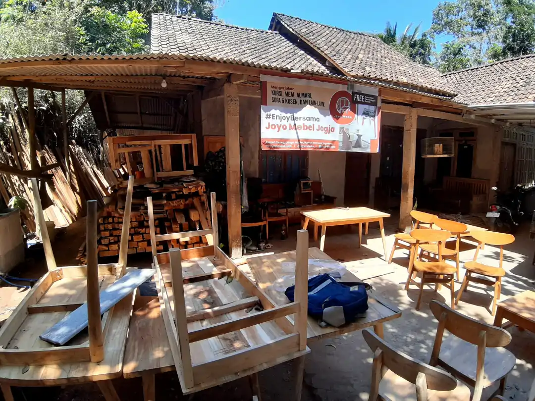 Workshop dengan furnitur kayu buatan tangan, tumpukan kayu, dan spanduk Joyo Mebel Jogja.