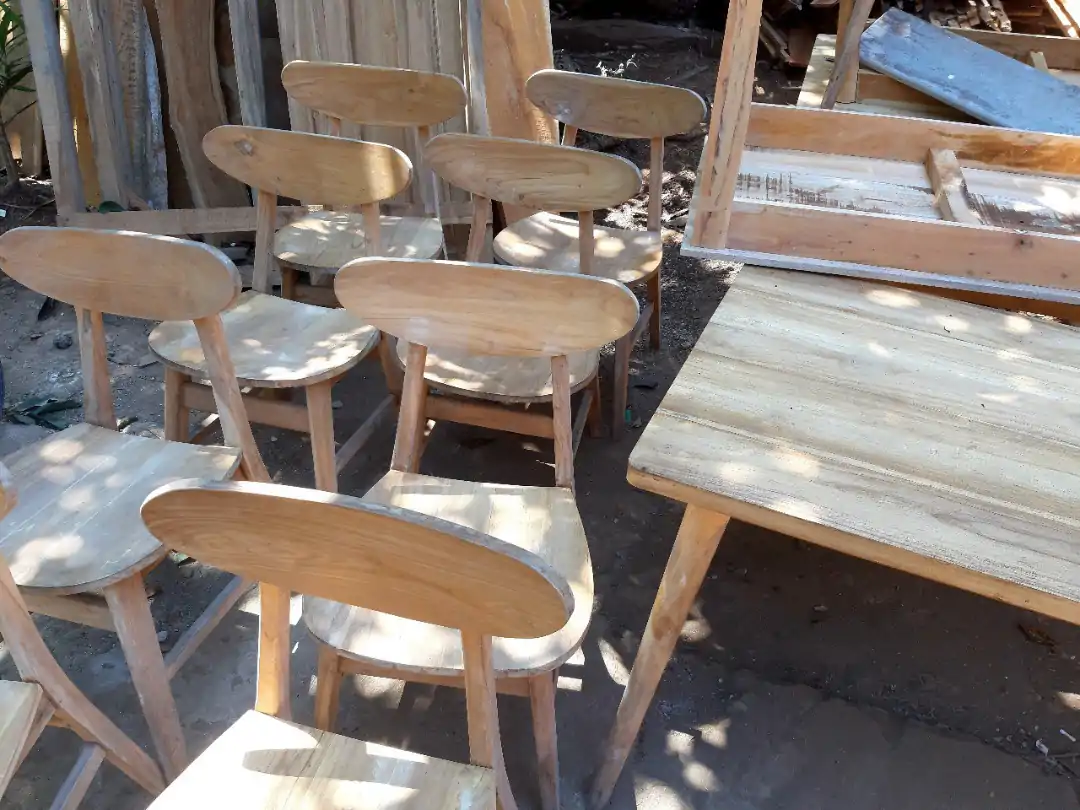 Satu set kursi kayu yang belum selesai dan meja di bengkel, dikelilingi oleh kayu dan peralatan pertukangan.